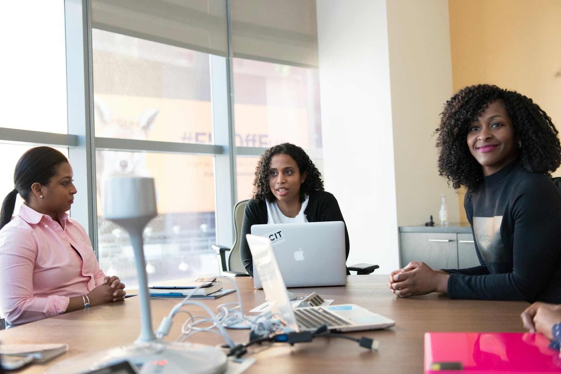 Two women in conversation at a modern boardroom table with a city view behind them.